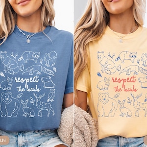 K&ouml;nnte beinhalten: Zwei T-Shirts, eines in Blau und eines in Gelb, jeweils mit einer wei&szlig;en Illustration verschiedener Tiere und dem Text "respect the locals". Die Tiere umfassen einen Fuchs, einen Waschb&auml;ren, ein Eichh&ouml;rnchen, ein Kaninchen, einen B&auml;ren, ein Reh und eine Eule.