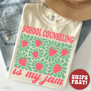 K&ouml;nnte beinhalten: Cremefarbenes T-Shirt mit dem Text "SCHOOL COUNSELING is my jam" in Rosa. Das Shirt zeigt ein Muster aus Erdbeeren und gr&uuml;nen floralen Designs. Ein roter Kreis mit "SHIPS FAST!" befindet sich unten rechts.