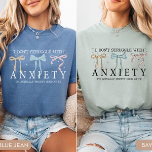 May include: Two women are wearing blue and green t-shirts with the text "I don't struggle with anxiety. I'm actually pretty good at it." and a graphic of three bows. The women are wearing blue jeans.