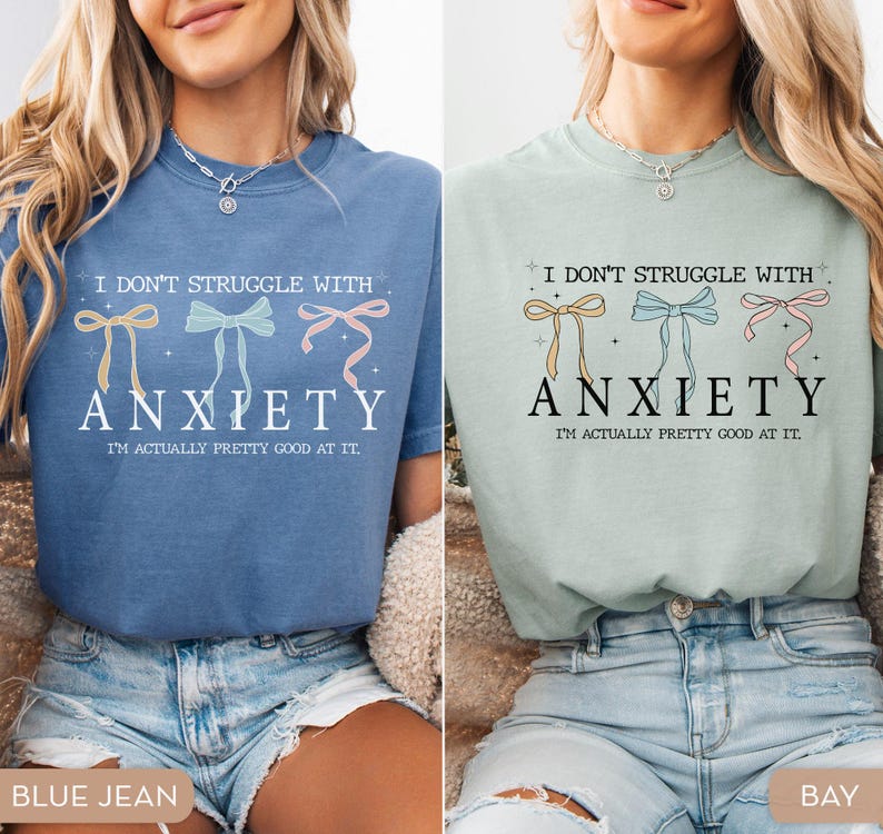 May include: Two women are wearing blue and green t-shirts with the text "I don't struggle with anxiety. I'm actually pretty good at it." and a graphic of three bows. The women are wearing blue jeans.