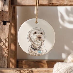 May include: A round white ceramic ornament featuring a detailed illustration of a cream-colored dog with curly fur. The dog wears a dark collar with a light blue bone-shaped tag. A gold string is attached to the top for hanging the ornament.