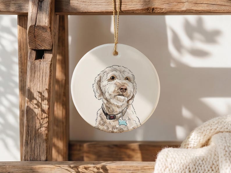 May include: A round white ceramic ornament featuring a detailed illustration of a cream-colored dog with curly fur. The dog wears a dark collar with a light blue bone-shaped tag. A gold string is attached to the top for hanging the ornament.