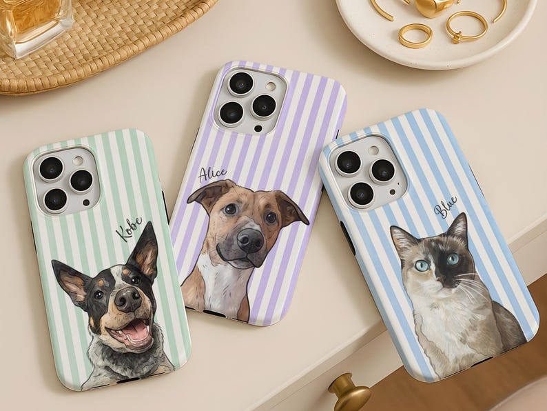 May include: Three smartphone cases featuring custom pet portraits. The left case has a mint green striped background with a dog named Kobe. The center case has a lavender striped background with a dog named Alice. The right case has a light blue striped background with a cat named Blue.
