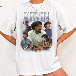 May include: A white short-sleeve t-shirt featuring a graphic print with the name KEVIN in large metallic-style letters at the top. The design includes five photographic portraits of a person with curly hair in various poses.