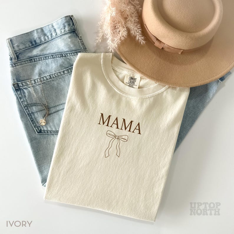 May include: Ivory t-shirt with the word "MAMA" and a brown bow printed on the front.