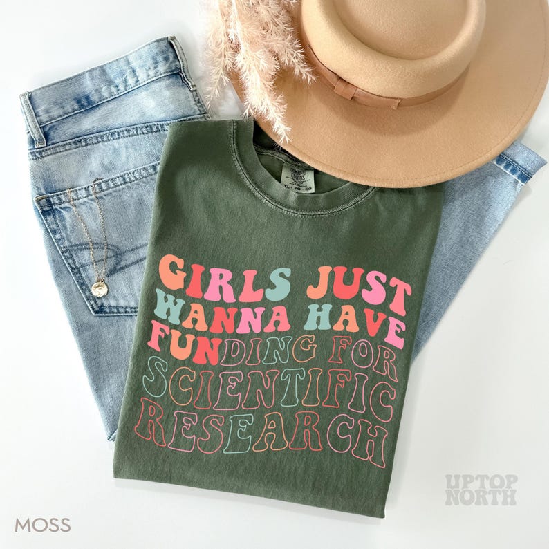 Pode incluir: Uma camiseta verde com o texto "Girls Just Wanna Have Funding For Scientific Research" em cores do arco-&iacute;ris.