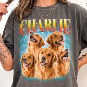 May include: A charcoal gray t-shirt featuring a graphic print of five golden retriever dogs arranged in a cluster. Above the dogs, the name CHARLIE is printed in large, bold, orange and yellow gradient letters against a blue background.