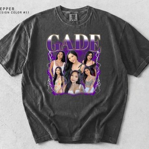 K&ouml;nnte beinhalten: Dunkelgraues T-Shirt mit einem Grafikdesign mit dem Namen "GADE" in lila und wei&szlig;, zusammen mit Bildern mehrerer Frauen. Das Shirt hat einen Vintage-Look.