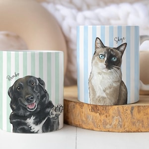 May include: Two white ceramic mugs featuring custom pet portraits. The left mug has a light green striped background with a black dog illustration labeled Rambo. The right mug has a light blue striped background with a cat illustration labeled Skye.