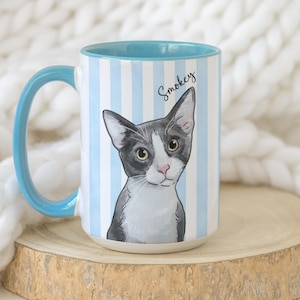 May include: A ceramic mug with a light blue and white vertical striped pattern. The mug features a graphic illustration of a grey and white cat with the name Smokey written in black script above the cat. The handle and interior are light blue.