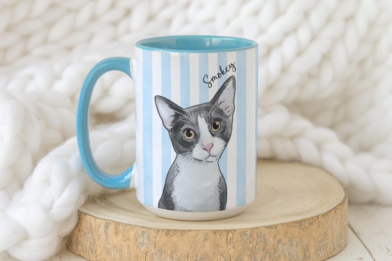 May include: A ceramic mug with a light blue and white vertical striped pattern. The mug features a graphic illustration of a grey and white cat with the name Smokey written in black script above the cat. The handle and interior are light blue.