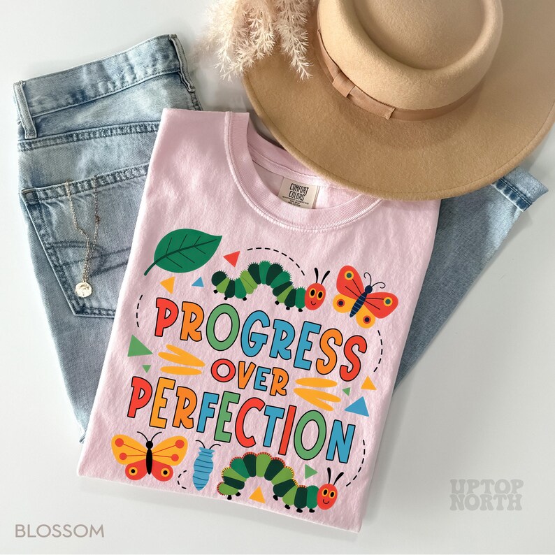 Op de afbeelding: Een lichtroze t-shirt met de tekst "PROGRESS OVER PERFECTION" in kleurrijke letters, versierd met cartoonrupsen, vlinders en geometrische vormen. Het shirt wordt gecombineerd met een lichtblauwe jeans en een beige hoed.
