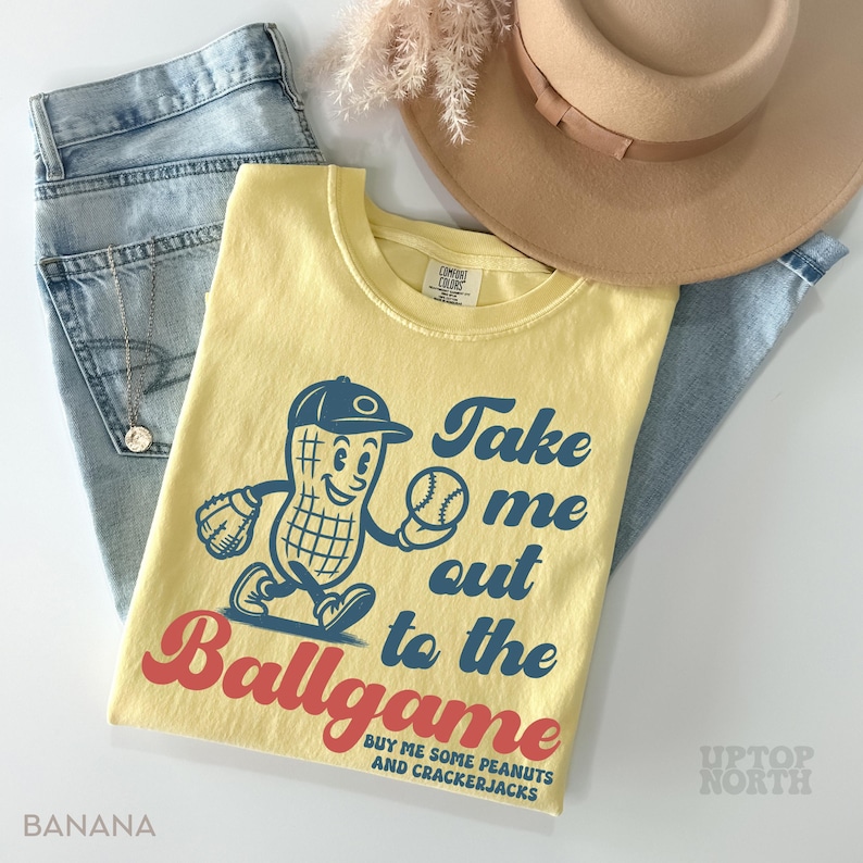 May include: A yellow t-shirt with a cartoon peanut wearing a baseball cap and holding a baseball. The text on the shirt reads "Take me out to the Ballgame Buy me some peanuts and crackerjacks".