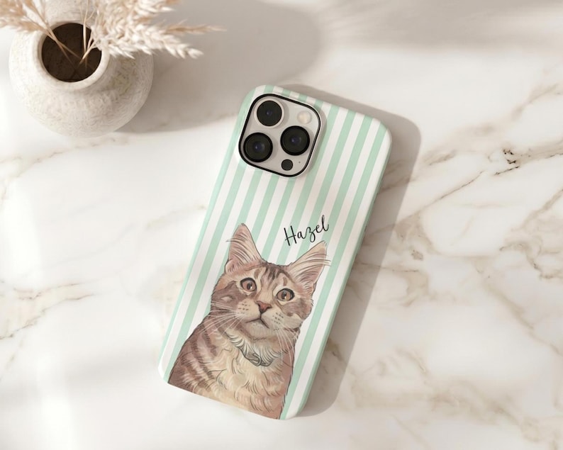 May include: A phone case featuring a cat illustration. The case has a mint green and white striped background. The cat is brown and tan with a white collar. The name "Hazel" is written in script on the case.