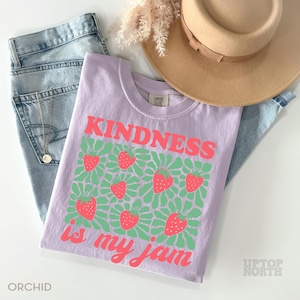 May include: A light purple t-shirt with the words "KINDNESS is my jam" in pink, along with a strawberry design. The shirt is paired with blue jeans and a tan hat. The word "ORCHID" is printed at the bottom left.