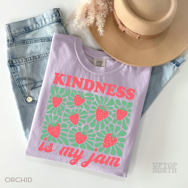 May include: A light purple t-shirt with the words "KINDNESS is my jam" in pink, along with a strawberry design. The shirt is paired with blue jeans and a tan hat. The word "ORCHID" is printed at the bottom left.