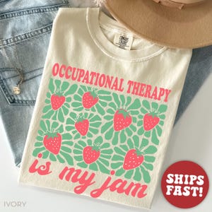 Op de afbeelding: Ivoor t-shirt met de tekst "OCCUPATIONAL THERAPY is my jam" in roze. Het shirt heeft een patroon van roze aardbeien op een groene achtergrond. Het shirt ligt op een blauwe jeans.