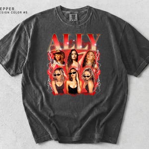 May include: A dark gray t-shirt featuring a graphic of the word "ALLY" in red, with a vintage-style design. The graphic includes images of six women. The shirt has a crew neck and short sleeves.