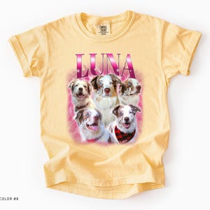 May include: A butter-colored short-sleeve t-shirt featuring a graphic print of five Australian Shepherd dogs. The word LUNA is printed in large, pink, gradient lettering above the dogs, which are set against a soft, pink, hazy background.
