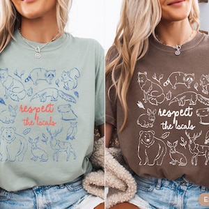 K&ouml;nnte beinhalten: Zwei T-Shirts, eines in Bay Green und das andere in Espresso-Braun, jeweils mit einer Grafik von Tieren und dem Text "respect the locals". Die Tiere umfassen einen B&auml;ren, ein Reh, ein Kaninchen und eine Eule.