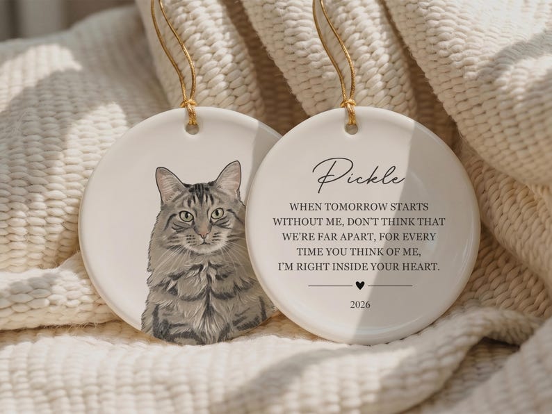 May include: Two round white ceramic ornaments with gold hanging strings. One features a detailed illustration of a grey tabby cat. The other displays the name Pickle, a memorial poem, a small heart icon, and the year 2026 in black text.