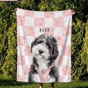 May include: A soft blanket featuring a pink and white checkered pattern with small pink paw prints. The center displays a portrait of a black and white dog wearing a pink harness, with the name NOVA printed in black capital letters above it.