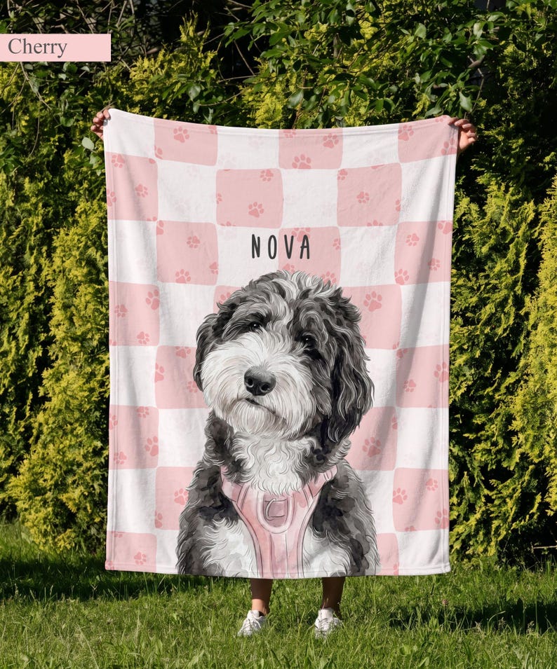 May include: A soft blanket featuring a pink and white checkered pattern with small pink paw prints. The center displays a portrait of a black and white dog wearing a pink harness, with the name NOVA printed in black capital letters above it.