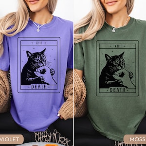 Op de afbeelding: Twee T-shirts, &eacute;&eacute;n violet en &eacute;&eacute;n mosgroen, elk met een zwarte afbeelding van een kat die een menselijke hand vasthoudt, omlijst door een tarotkaartontwerp. De kaart leest "XIII" bovenaan en "DEATH" onderaan.