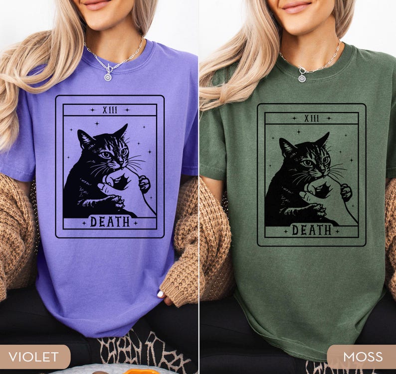 Op de afbeelding: Twee T-shirts, &eacute;&eacute;n violet en &eacute;&eacute;n mosgroen, elk met een zwarte afbeelding van een kat die een menselijke hand vasthoudt, omlijst door een tarotkaartontwerp. De kaart leest "XIII" bovenaan en "DEATH" onderaan.