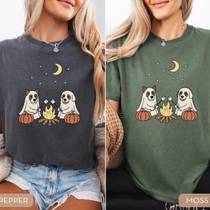 May include: Two t-shirts, one pepper and one moss green, each featuring a graphic of two dogs roasting marshmallows over a campfire, with pumpkins, a crescent moon, and stars. The shirts are short-sleeved.