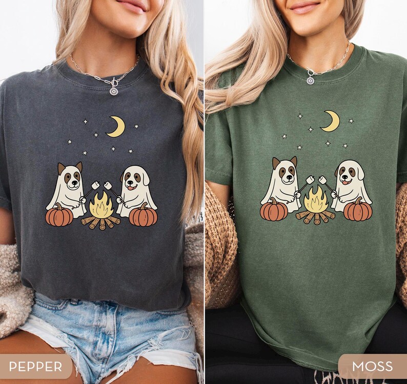 May include: Two t-shirts, one pepper and one moss green, each featuring a graphic of two dogs roasting marshmallows over a campfire, with pumpkins, a crescent moon, and stars. The shirts are short-sleeved.