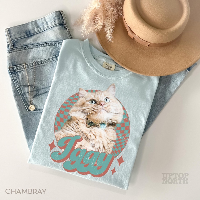 May include: Light blue t-shirt featuring a cat graphic with the name "Iggy" in a retro font. The shirt is paired with light wash denim jeans and a tan hat. A gold necklace is visible.