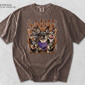 Pu&ograve; includere: T-shirt color espresso con il nome "SADIE" in un carattere marrone invecchiato. La maglietta presenta una grafica di cinque cani, uno dei quali indossa una bandana viola. Il design include accenti di fulmini.