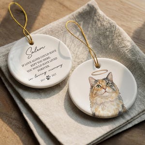 May include: Two round white ceramic ornaments with gold hanging strings. One features the text Salem, If love alone could have kept you here, you would have lived forever, in loving memory, 2011 paw print 2025. The other shows a calico cat with a halo.