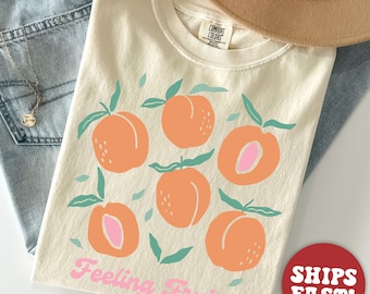 Puede incluir: Una camiseta de color crema con un gráfico de melocotones y el texto "Feeling Fruity" en rosa. La camiseta está hecha de un material suave y cómodo.