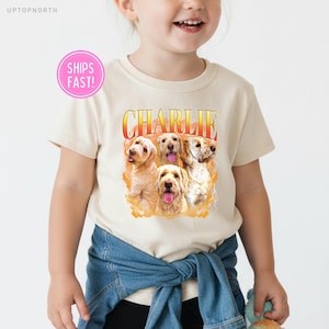 May include: A cream-colored short-sleeve t-shirt featuring a graphic print of four golden-colored dogs. Above the dogs, the name CHARLIE is printed in a bold, orange-to-yellow gradient font. The graphic is set against a soft orange background.