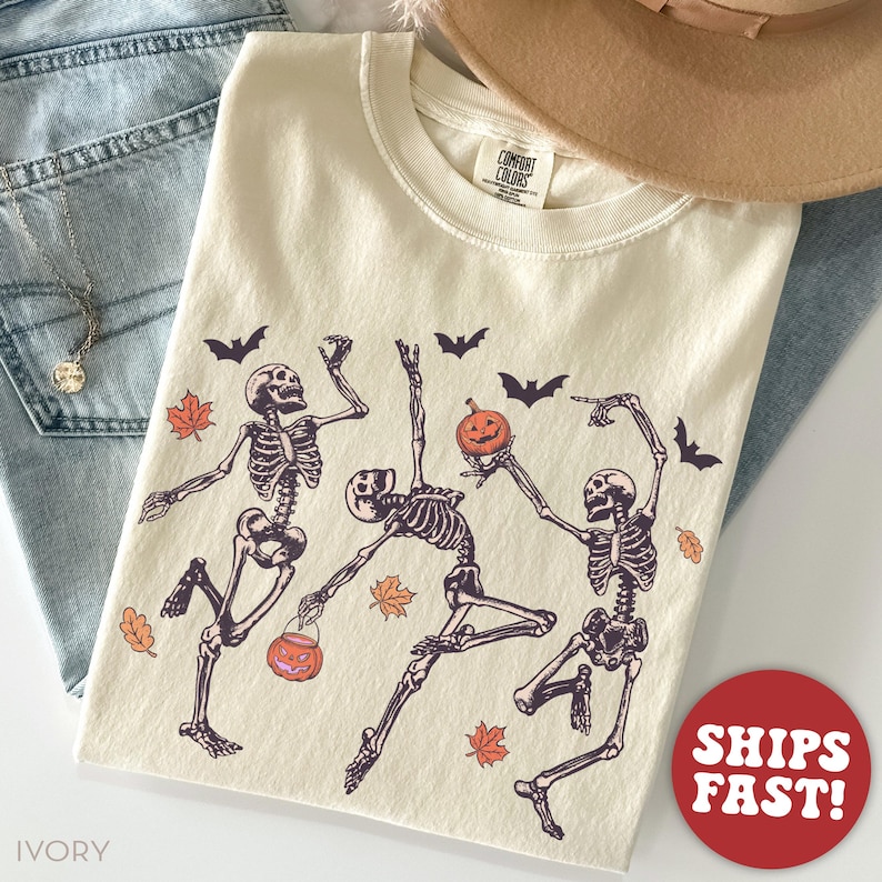 K&ouml;nnte beinhalten: Ein elfenbeinfarbenes T-Shirt mit einem Grafikdruck von drei tanzenden Skeletten, die einen K&uuml;rbis und einen K&uuml;rbiseimer halten, umgeben von schwarzen Flederm&auml;usen und orangefarbenen Herbstbl&auml;ttern. Das Shirt hat ein Comfort Colors Etikett am Hals.