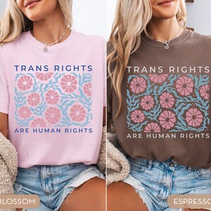 May include: Two t-shirts with the text "Trans Rights Are Human Rights" in blue and pink lettering. The shirts have a floral design in pink and blue. The shirt on the left is pink and the shirt on the right is brown. The shirts are labeled "Blossom" and "Espresso".