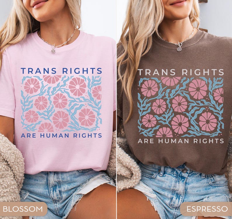 May include: Two t-shirts with the text "Trans Rights Are Human Rights" in blue and pink lettering. The shirts have a floral design in pink and blue. The shirt on the left is pink and the shirt on the right is brown. The shirts are labeled "Blossom" and "Espresso".