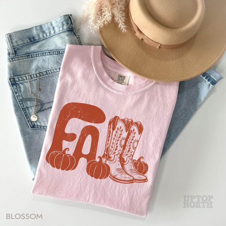 May include: A light pink t-shirt with the word "FALL" in orange, featuring pumpkins and cowboy boots. The shirt is paired with light blue jeans and a tan hat, creating a fall-themed aesthetic. The word "BLOSSOM" is printed at the bottom left.