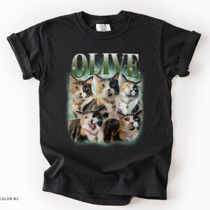 May include: A black short-sleeve t-shirt featuring a graphic print of five calico cats arranged in a cluster. Above the cats, the word OLIVE is printed in a large, bold, green-tinted font. The cats wear various collars with small charms.