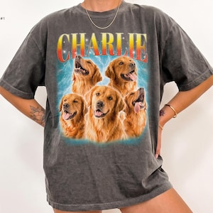 May include: A charcoal gray short-sleeve t-shirt featuring a graphic print of four golden retriever dogs. Above the dogs, the name CHARLIE is printed in a bold, red-to-yellow gradient font. The background of the graphic is a light blue.