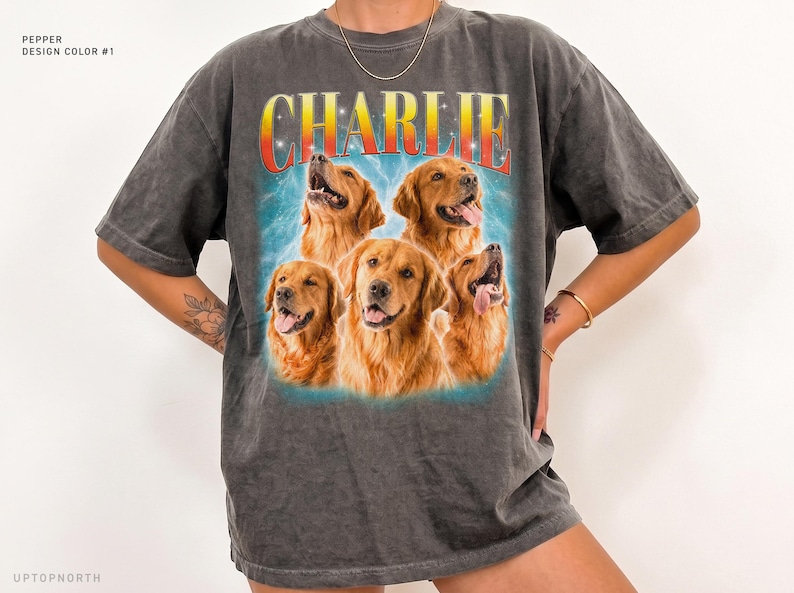 May include: A charcoal gray short-sleeve t-shirt featuring a graphic print of four golden retriever dogs. Above the dogs, the name CHARLIE is printed in a bold, red-to-yellow gradient font. The background of the graphic is a light blue.