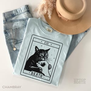 Op de afbeelding: Een lichtblauw t-shirt met een zwart-witte katten tarotkaart afbeelding met het woord "DEATH". Het shirt ligt op een lichtblauwe jeans, een gouden ketting en een beige hoed. Er zijn ook gedroogde bloemen te zien.