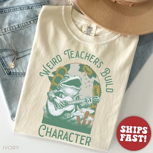 May include: Ivory-colored t-shirt with a graphic of a frog playing a guitar, surrounded by sunflowers and mushrooms. The text "Weird Teachers Build Character" is arched around the frog. A red circle with "Ships Fast!" is in the lower right.