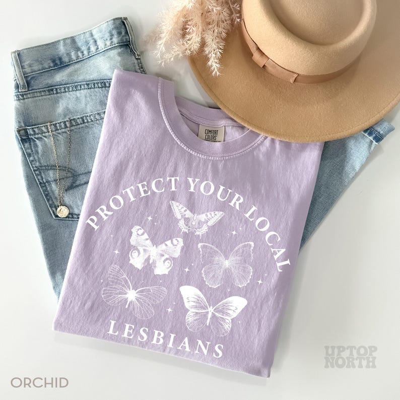 May include: A light purple t-shirt with the text "PROTECT YOUR LOCAL LESBIANS" and butterfly graphics. The shirt is paired with light blue jeans, a tan hat, and a gold necklace. The word "ORCHID" is printed at the bottom left.
