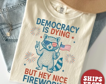 Op de afbeelding: Ivoor kleurig t-shirt met een afbeelding van een wasbeer met een zonnebril met een Amerikaans vlagontwerp, die een kleine Amerikaanse vlag vasthoudt en een vredesteken geeft. De tekst luidt "DEMOCRACY IS DYING BUT HEY NICE FIREWORKS."