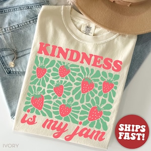 May include: Ivory-colored t-shirt with the words "KINDNESS is my jam" in pink. The shirt features a pattern of strawberries and green leaves. A brown hat and blue jeans are in the background.