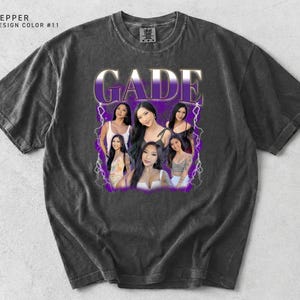 May include: Dark gray t-shirt with a graphic design featuring the name "GADE" in a purple and white font, along with images of several women. The shirt has a vintage, worn look.