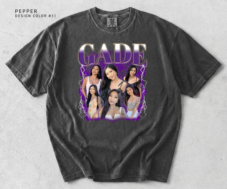 May include: Dark gray t-shirt with a graphic design featuring the name "GADE" in a purple and white font, along with images of several women. The shirt has a vintage, worn look.